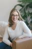 Kelley Nay sits relaxed on a tan leather chair, wearing glasses, a cream sweater, and a gold necklace. She rests one arm on the chair and appears calm and approachable. The softly lit room features a white wall, a plush blanket, and green leafy plants in the background, creating a warm and welcoming atmosphere.