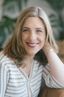 Kelley Nay smiles warmly while seated, wearing a patterned dress. Her posture is relaxed and inviting. The softly blurred background features neutral tones and hints of greenery, creating a calm and welcoming atmosphere.