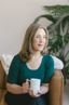 Kelley Nay sits thoughtfully in a green, sunlit space. She wears glasses and a soft sweater, appearing warm and approachable. The background features plants and calming decor creating a peaceful, inviting atmosphere.